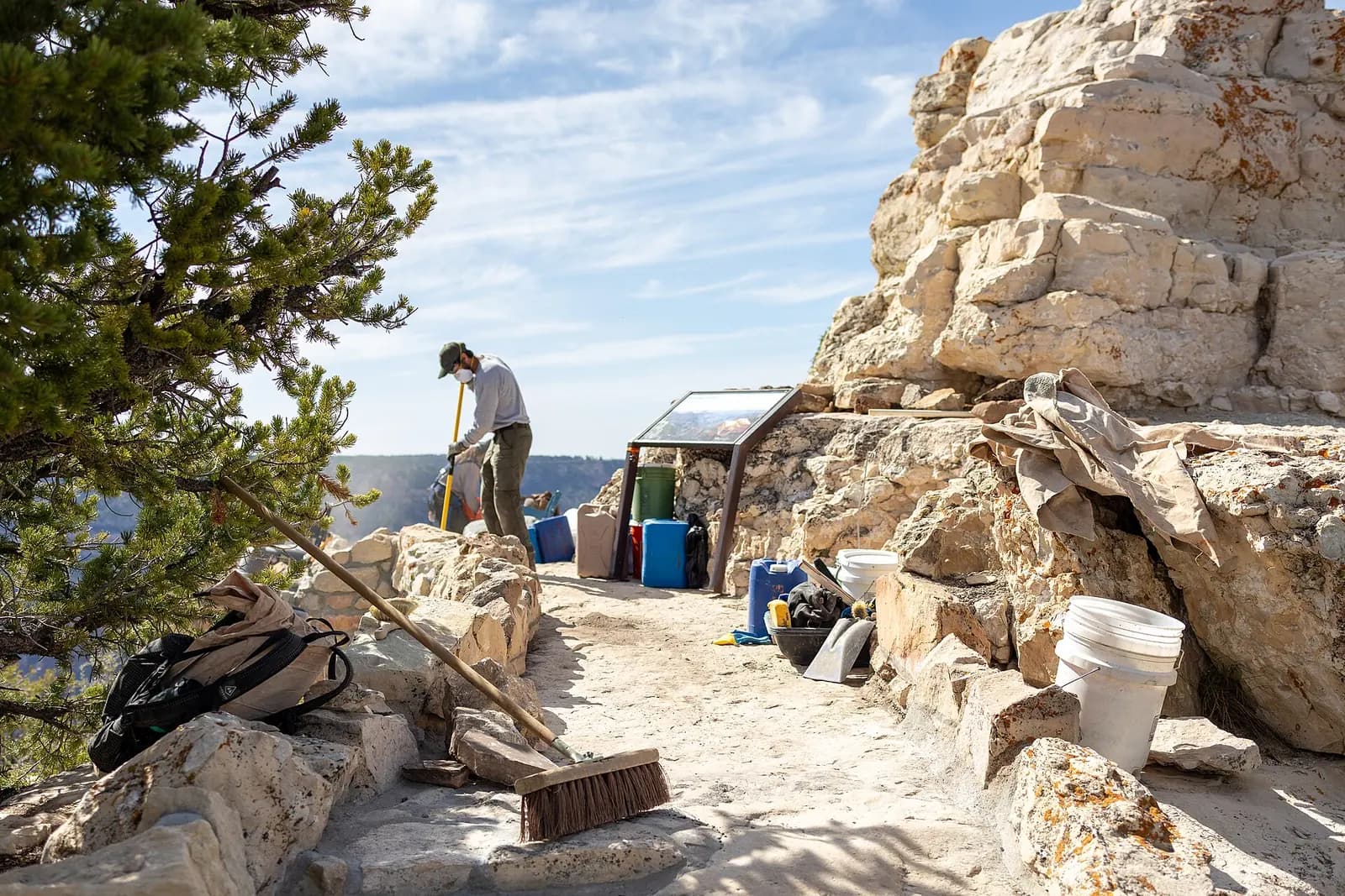 A trail worker raking fresh tread at a canyon-rim work site, surrounded by buckets, hand tools and stacked stone.