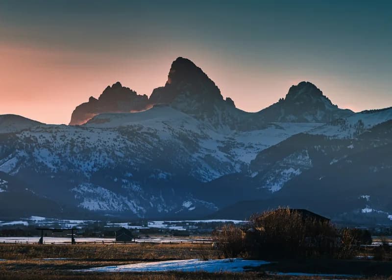 The snow-covered Teton range at first light seen across open Idaho farmland, with a barn and bare trees in the foreground.
