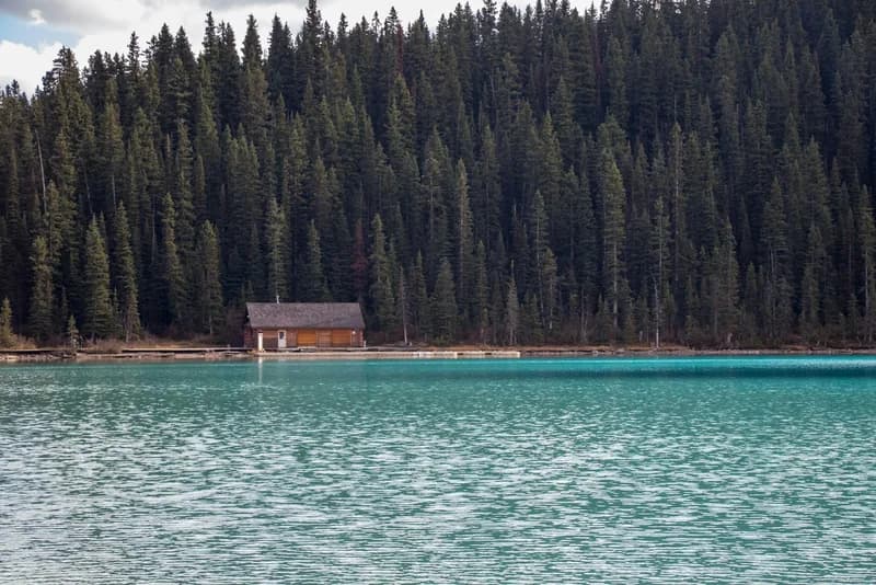 A small log boathouse on the shore of a turquoise glacial lake, backed by a dense wall of evergreen forest.