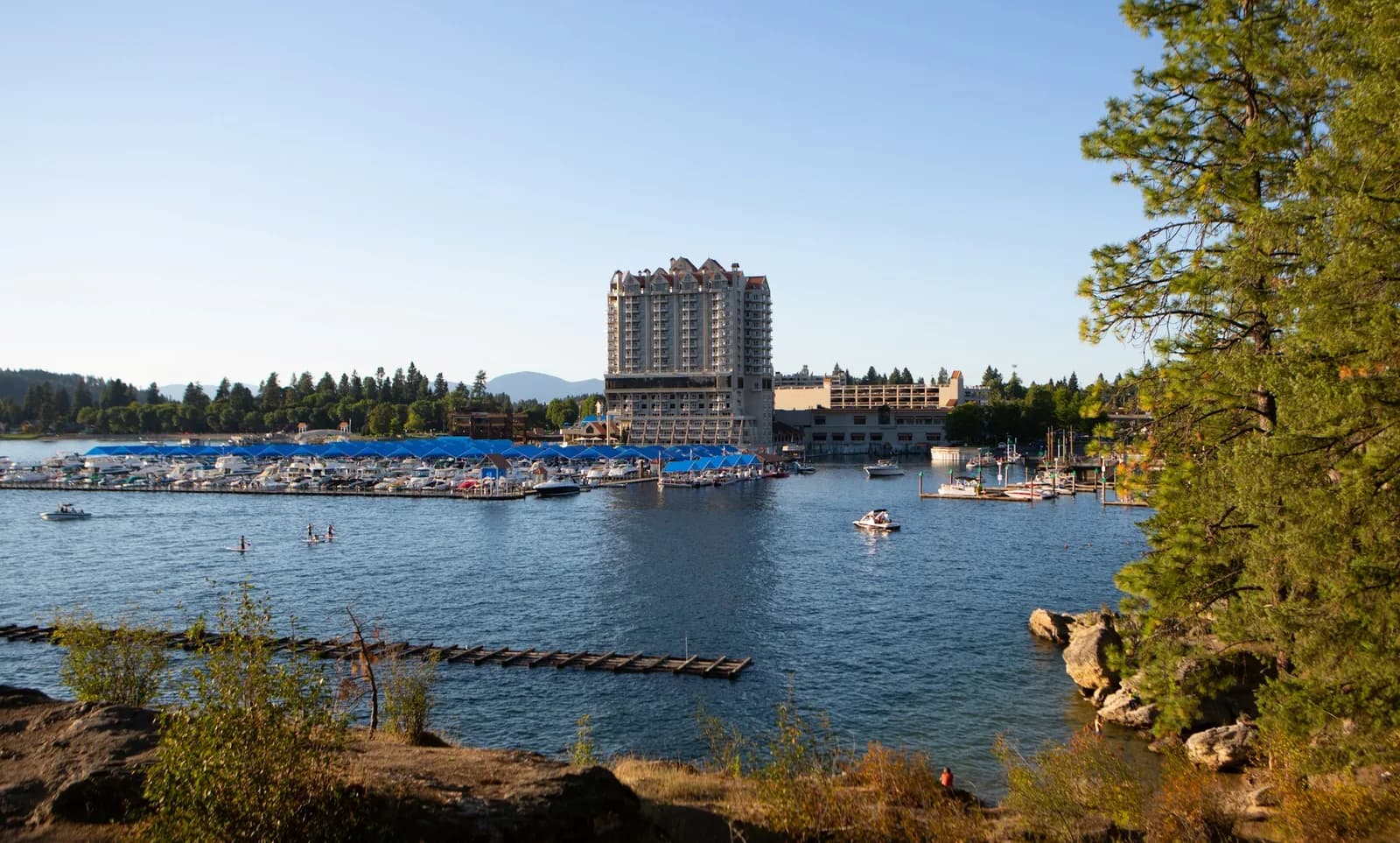 The Coeur d'Alene Resort tower rising above a marina full of moored boats, with a pine-covered shoreline and calm blue water in the foreground.